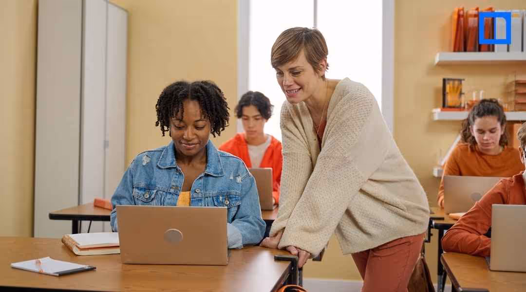 A female teacher is leaning over and smiling at a female student of color who is working on a laptop in a classroom. Other students are also visible, working at their desks with laptops.