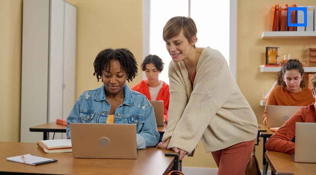 A female teacher is leaning over and smiling at a female student of color who is working on a laptop in a classroom. Other students are also visible, working at their desks with laptops.