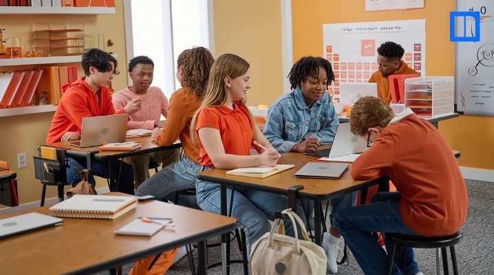 A diverse group of students in a classroom sitting at desks and collaborating on an activity. They are engaged with their laptops, notebooks, and discussions. The room has science-themed decor, including a periodic table on the back wall.