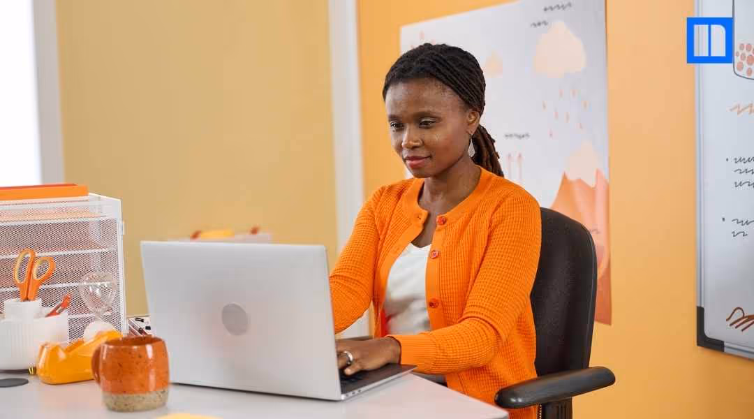 A female teacher with braided hair is smiling while looking at her laptop screen. She is wearing a white top and an orange cardigan and is seated at a desk in a classroom.