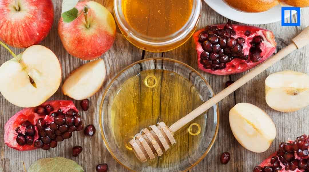 An overhead view of a wooden table set with traditional Rosh Hashanah foods. There are whole and sliced apples, pomegranates, and bowls of honey, with a honey dipper in one of the bowls.