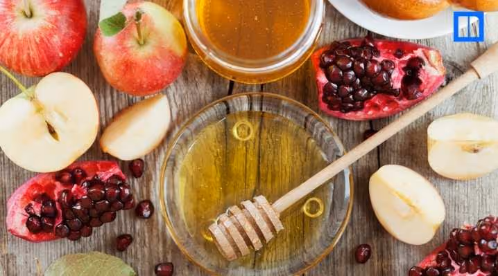 An overhead view of a wooden table set with traditional Rosh Hashanah foods. There are whole and sliced apples, pomegranates, and bowls of honey, with a honey dipper in one of the bowls.