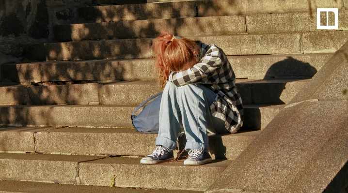 A young person with red hair is sitting alone on concrete stairs with their head down and knees pulled up, looking sad and isolated.