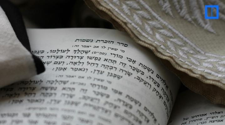 A close-up, overhead view of an open prayer book with Hebrew text. A white tallit (prayer shawl) with embroidered trim is visible in the top right corner.