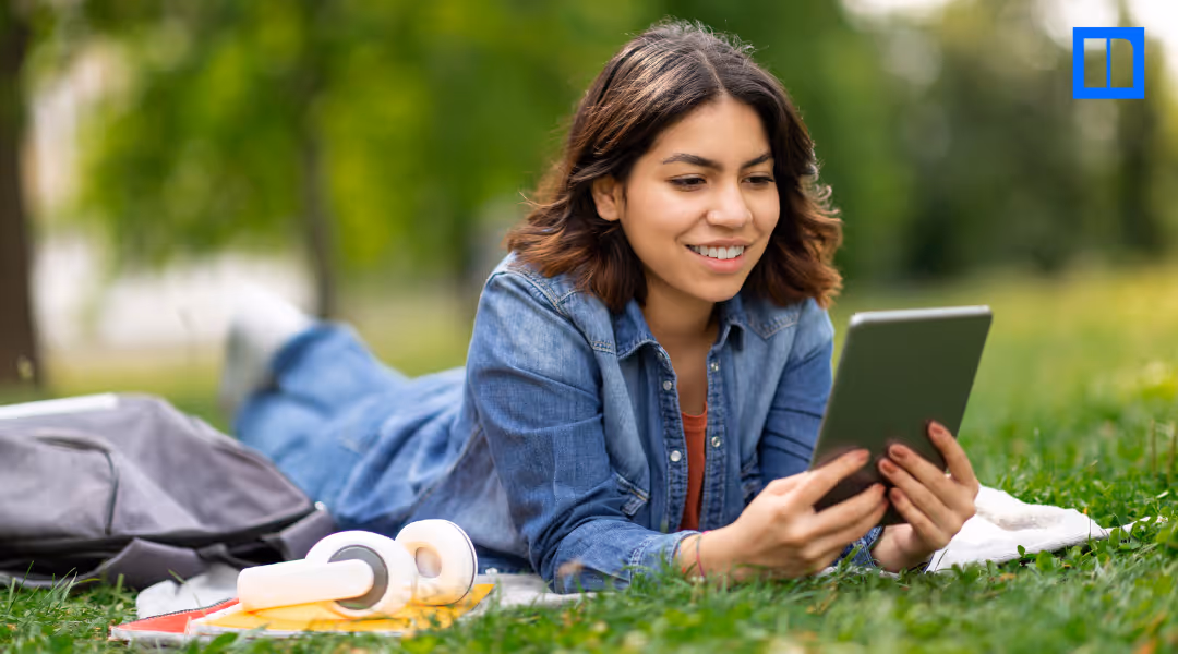 A smiling female student is lying on her stomach in a grassy park, looking at a tablet. A backpack and headphones are next to her on a blanket.