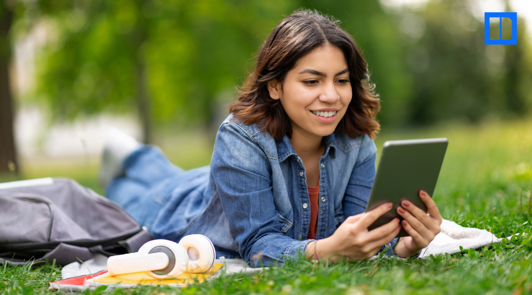 A smiling female student is lying on her stomach in a grassy park, looking at a tablet. A backpack and headphones are next to her on a blanket.