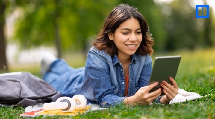 A smiling female student is lying on her stomach in a grassy park, looking at a tablet. A backpack and headphones are next to her on a blanket.