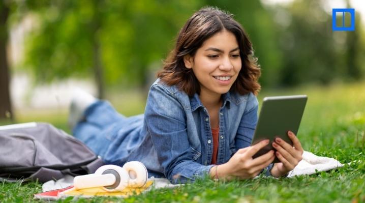 A smiling female student is lying on her stomach in a grassy park, looking at a tablet. A backpack and headphones are next to her on a blanket.