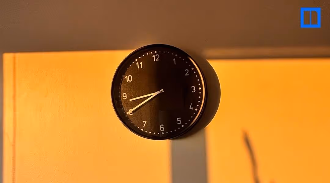 A black and white circular clock on a light-colored wall. The clock's hands are at 8:40, with a long shadow cast across the wall from a light source, creating a warm, golden hue.