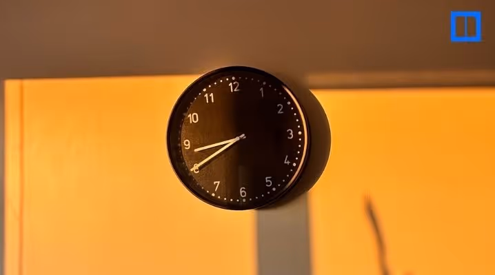 A black and white circular clock on a light-colored wall. The clock's hands are at 8:40, with a long shadow cast across the wall from a light source, creating a warm, golden hue.