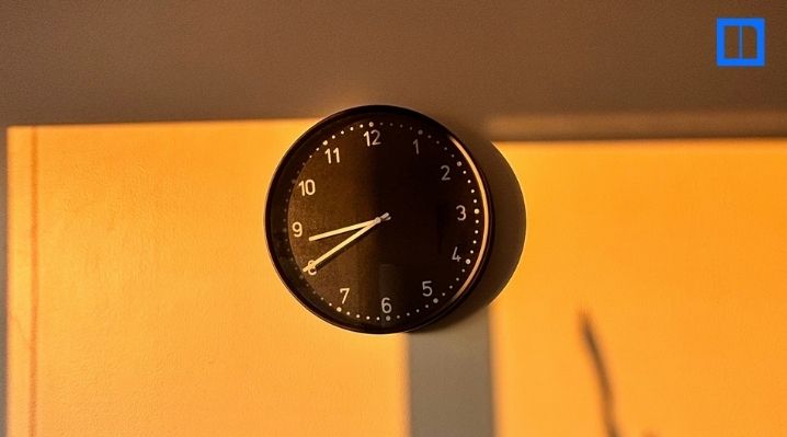 A black and white circular clock on a light-colored wall. The clock's hands are at 8:40, with a long shadow cast across the wall from a light source, creating a warm, golden hue.