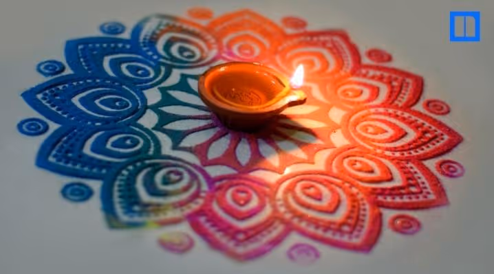 A close-up, overhead view of a lit clay oil lamp (diya) sitting in the center of a beautiful, circular rangoli design made of colorful powders. The flame casts a warm glow on the intricate pattern.