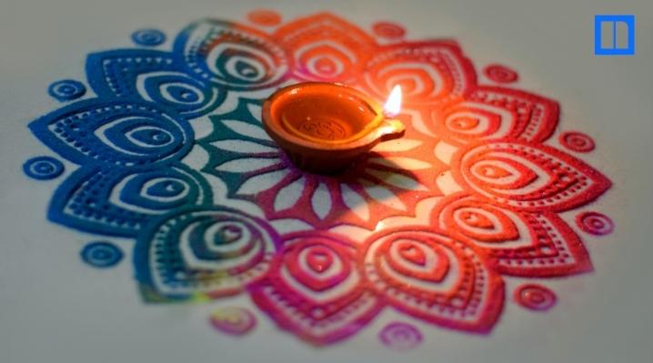 A close-up, overhead view of a lit clay oil lamp (diya) sitting in the center of a beautiful, circular rangoli design made of colorful powders. The flame casts a warm glow on the intricate pattern.