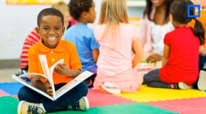 A young boy in an orange polo shirt smiles while holding a book during a classroom reading circle. Other students sit in the background on a colorful alphabet rug.