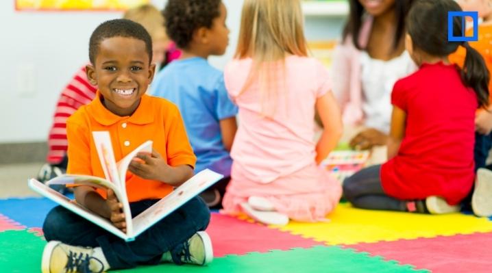 A young boy in an orange polo shirt smiles while holding a book during a classroom reading circle. Other students sit in the background on a colorful alphabet rug.
