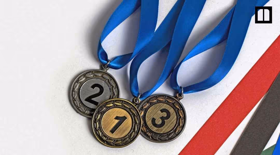 Close-up of gold, silver, and bronze Olympic medals numbered 1, 2, and 3 with blue ribbons, laid over decorative green, red, and yellow ribbons.