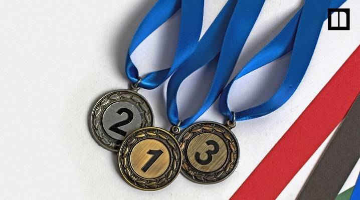 Close-up of gold, silver, and bronze Olympic medals numbered 1, 2, and 3 with blue ribbons, laid over decorative green, red, and yellow ribbons.