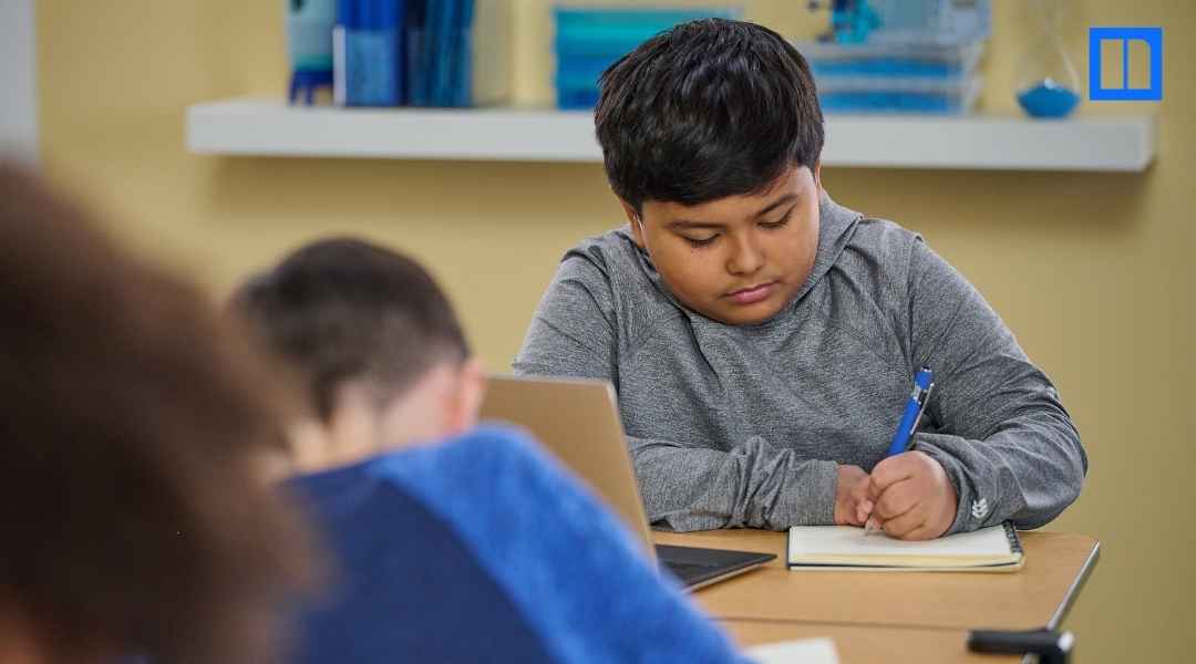 Close-up of a student in a grey hoodie writing in a notebook at a school desk during a classroom lesson, used as a hero image for a Newsela blog post about test preparation.