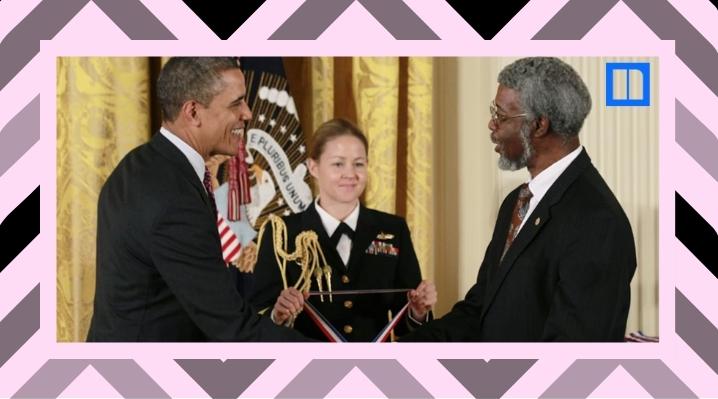 President Barack Obama smiling and shaking hands with theoretical physicist Dr. Sylvester James Gates Jr. during a White House ceremony. A military aide stands between them holding a medal.