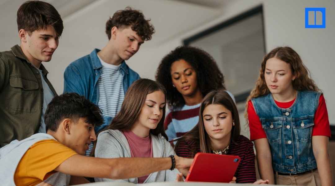 A wide-angle photo of six diverse teenage students leaning in to look at a red tablet together in a bright, modern classroom. The Newsela logo is visible in the top right corner.