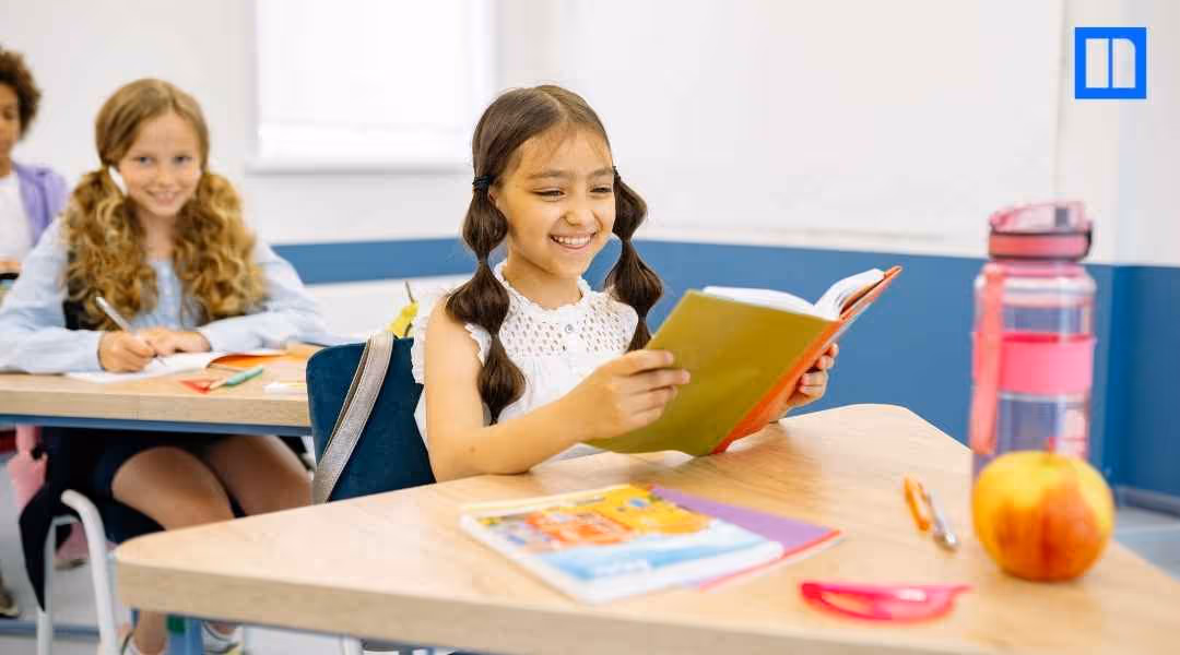 A young girl smiling while reading at her desk in a bright, modern elementary classroom with a blurred student in the background.