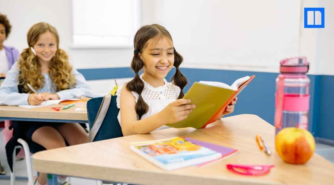 A young girl smiling while reading at her desk in a bright, modern elementary classroom with a blurred student in the background.