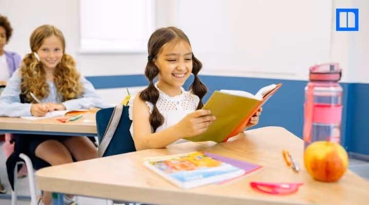 A young girl smiling while reading at her desk in a bright, modern elementary classroom with a blurred student in the background.