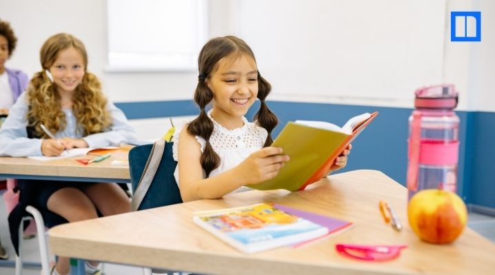 A young girl smiling while reading at her desk in a bright, modern elementary classroom with a blurred student in the background.