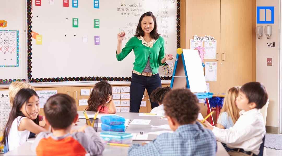 A female teacher in a green cardigan stands at the front of a bright elementary classroom, smiling and gesturing while teaching a group of young students sitting at a table.