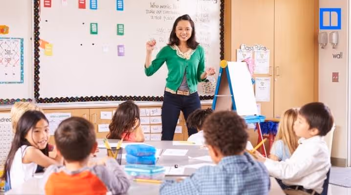 A female teacher in a green cardigan stands at the front of a bright elementary classroom, smiling and gesturing while teaching a group of young students sitting at a table.