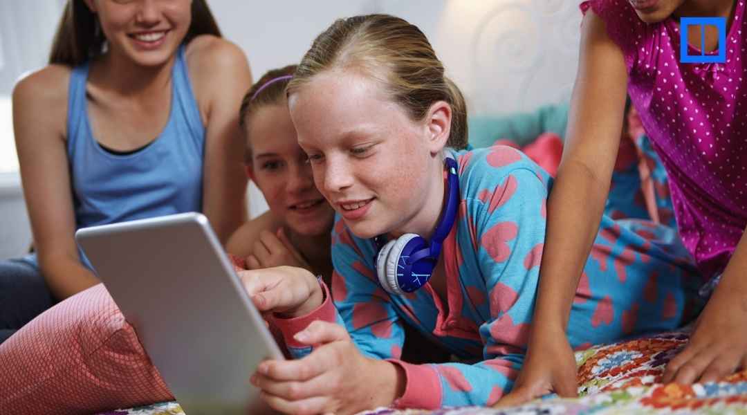 A group of diverse middle school students huddled together laughing while looking at a digital tablet. One girl in the center wears blue headphones around her neck.