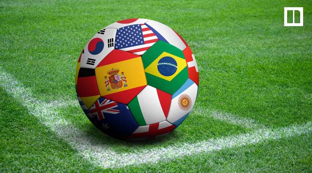 A soccer ball featuring various international flags—including Brazil, USA, Spain, and South Korea—sitting on the white corner line of a green grass soccer field.