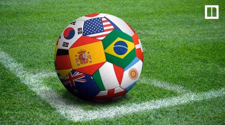 A soccer ball featuring various international flags—including Brazil, USA, Spain, and South Korea—sitting on the white corner line of a green grass soccer field.