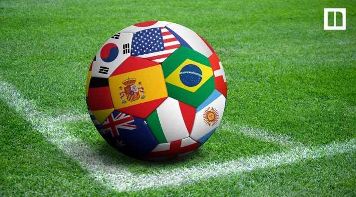 A soccer ball featuring various international flags—including Brazil, USA, Spain, and South Korea—sitting on the white corner line of a green grass soccer field.