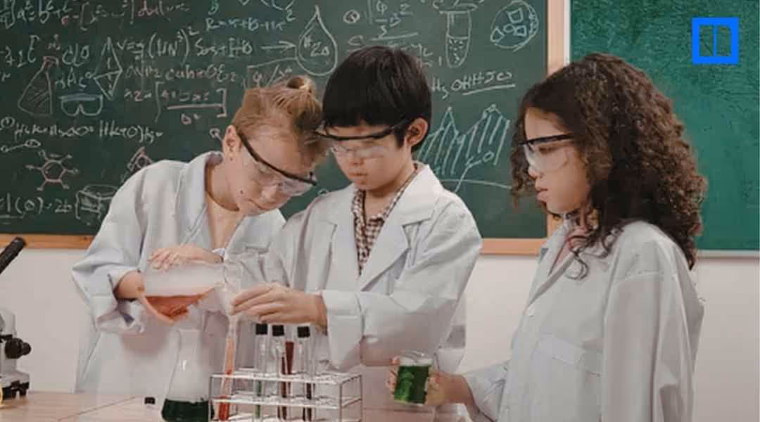 Three students wearing lab coats and safety goggles conduct a science experiment with test tubes and laboratory equipment in a classroom.