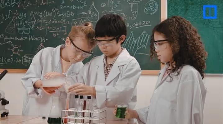 Three students wearing lab coats and safety goggles conduct a science experiment with test tubes and laboratory equipment in a classroom.