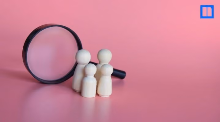Four wooden figures in front of a magnifying glass on a pink background.