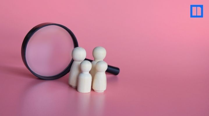 Four wooden figures in front of a magnifying glass on a pink background.