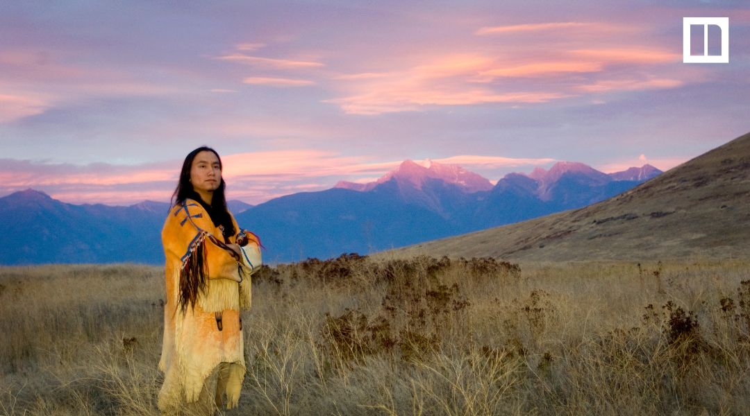 Person wearing a yellow fringed garment in a grassy mountain landscape at sunset, featured in a Native American Heritage Month social studies blog.