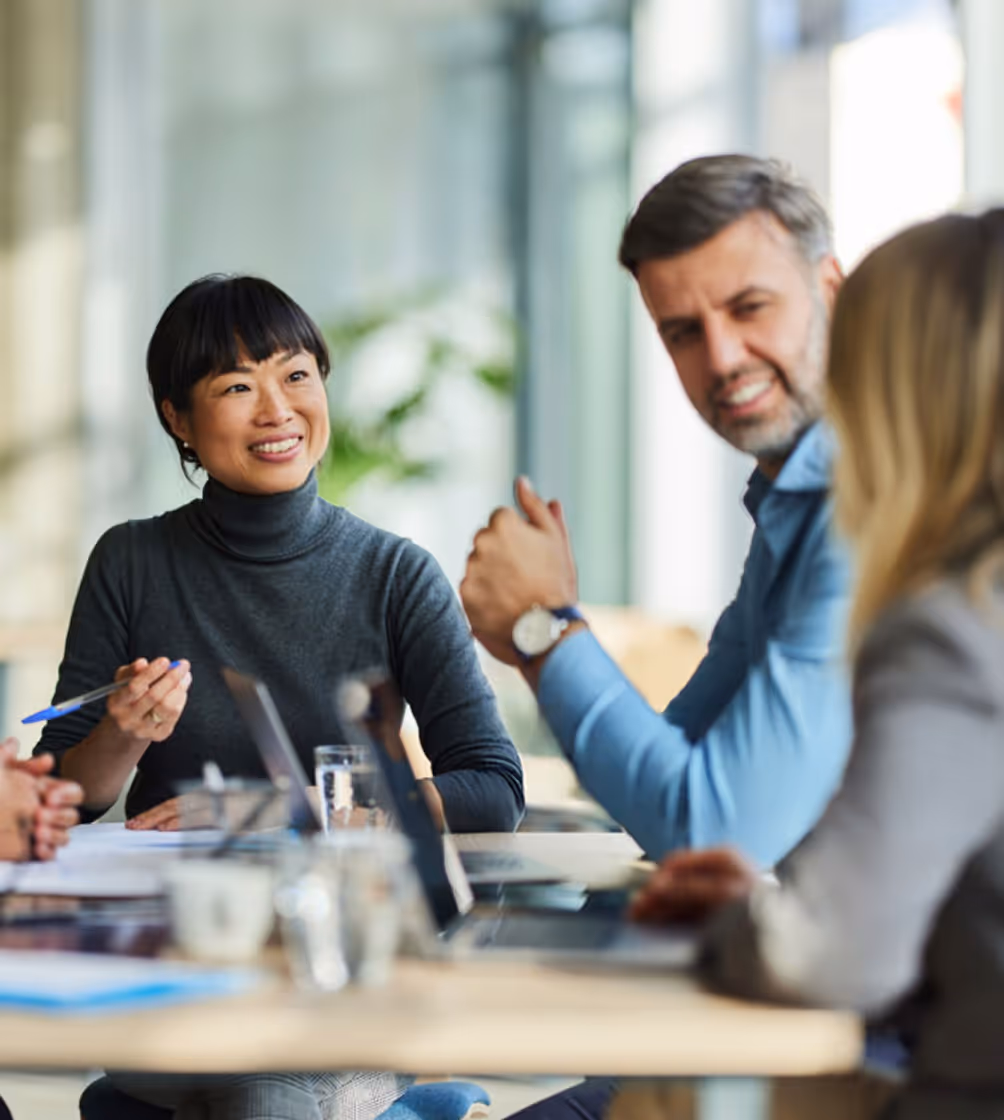 Three professionals engaged in a meeting around a table with laptops and documents, one woman smiling and holding a pen.