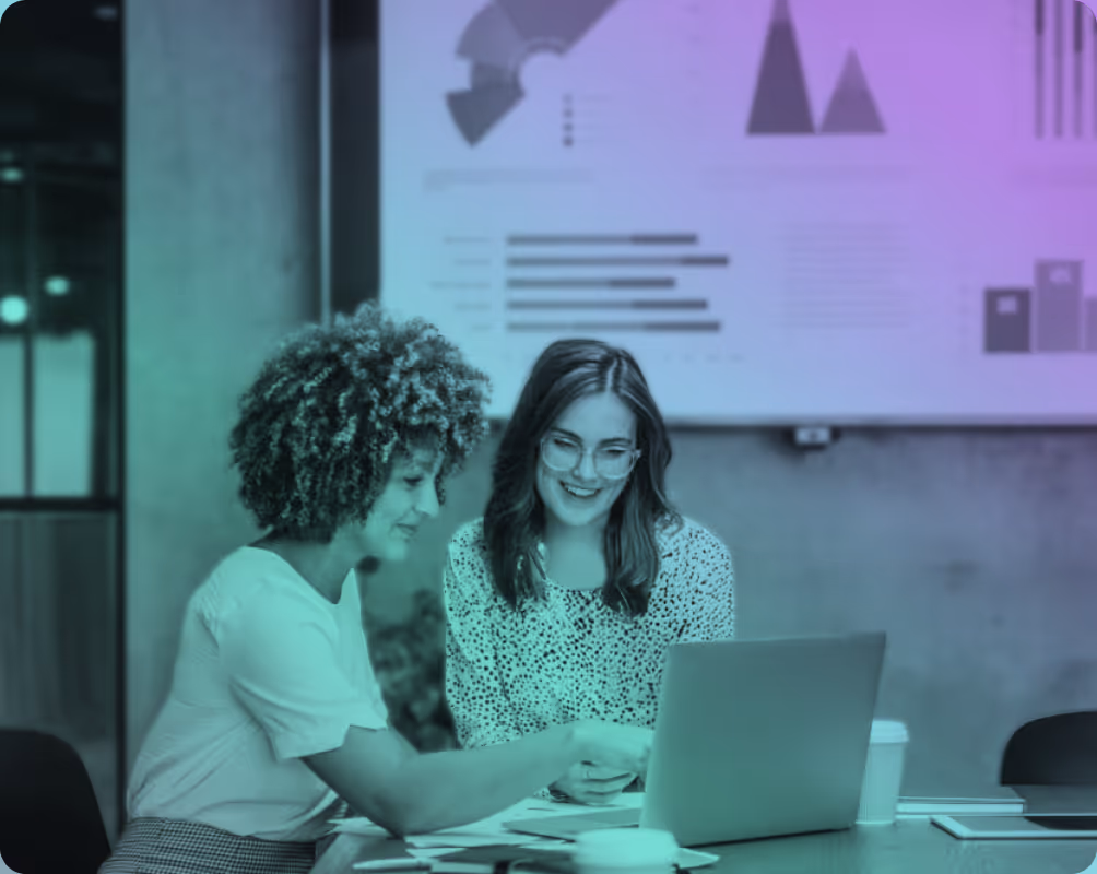 Two women seated at a table working together on a laptop with charts displayed on a screen behind them.