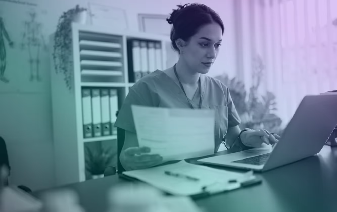 Medical professional in scrubs reviewing documents while working on a laptop at a desk in an office.
