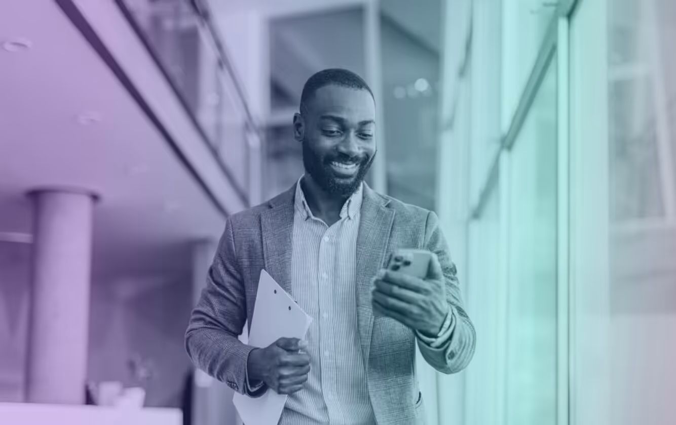 Smiling businessman holding folder in one hand and looking at smartphone in the other while walking indoors.