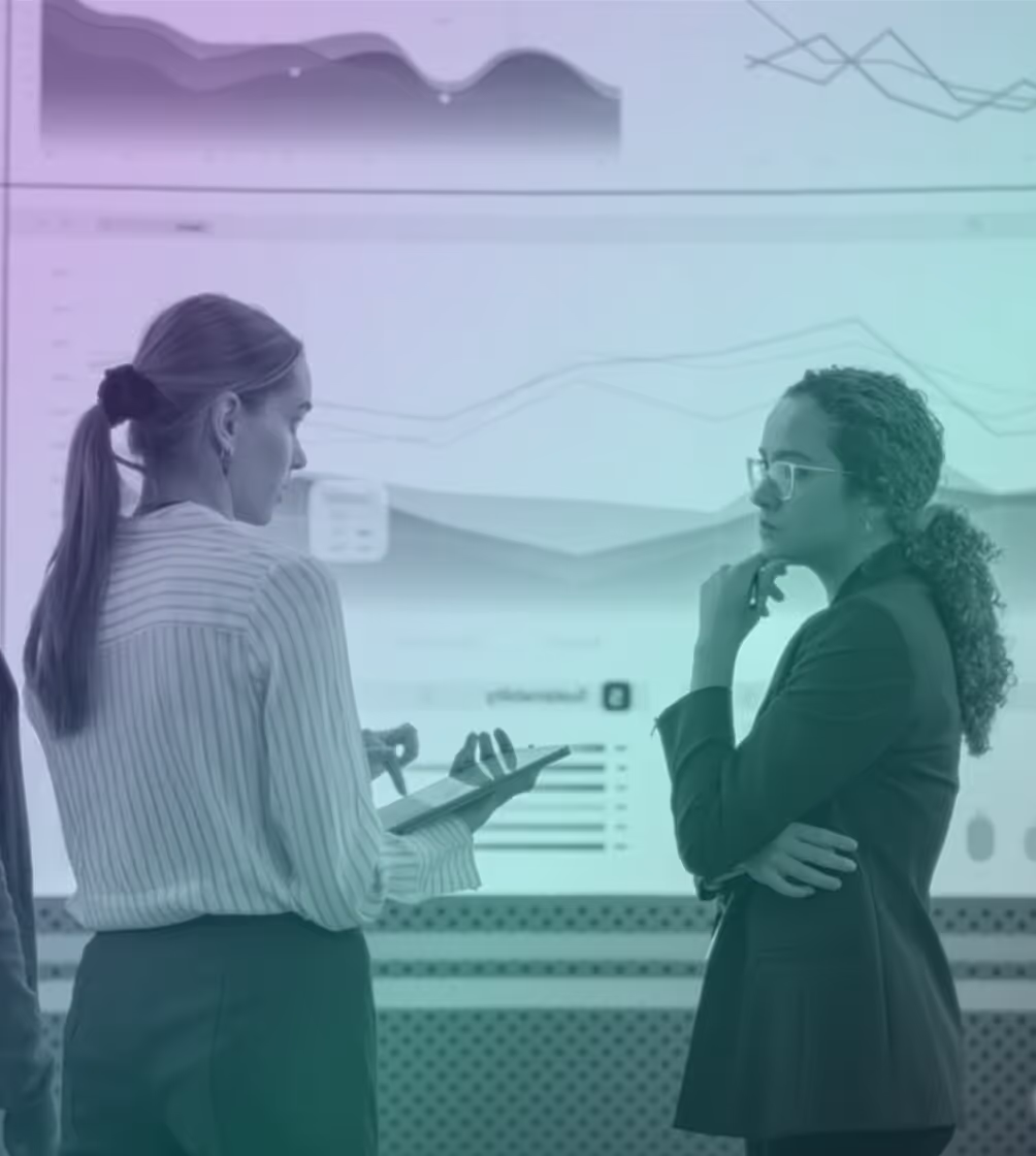 Two women discussing data charts displayed on a large screen, one holding a tablet and the other with a thoughtful expression.
