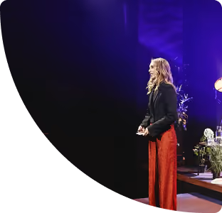 Vrouw staat lachend op een podium met rode broek en zwart jasje, omringd door planten en sfeerverlichting.