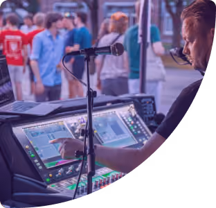 Een geluidsoperator bedient een digitale mengpaneel tijdens een buitenevenement met mensen op de achtergrond.
