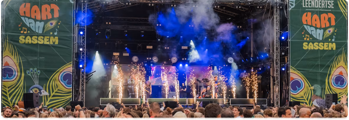 Een drukbezocht openluchtconcert met een podium waarop shows worden uitgevoerd met vuurfonteinen en rook, geflankeerd door groene banners met de tekst Hart Van Sassem en kleurige pauwenveren, terwijl het publiek op de voorgrond geniet.