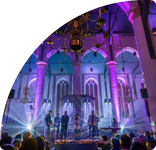 Muzikanten op een podium in een kerk met hoge zuilen verlicht door paarse en blauwe verlichting, met een kroonluchter aan het plafond en publiek op de voorgrond.