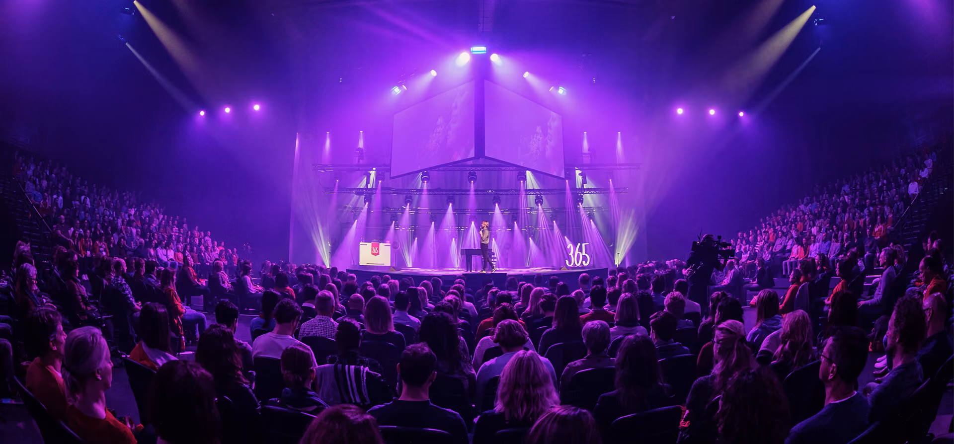 Publiek in een theaterzaal kijkt naar een spreker op een verlicht podium met paarse en gele spotlights en grote schermen erboven.