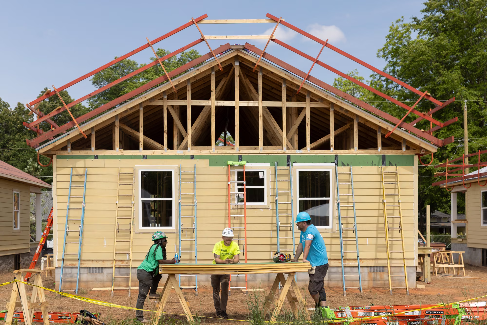 Three builders in hard hats talk across a sawhorse table in front of a framed Atlanta Habitat home